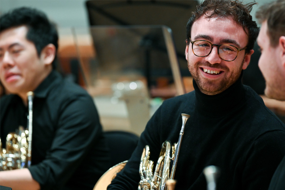 A male student, wearing a black shirt, smiling and chatting to other students, all sitting down and holding their French horn, in a rehearsal setting.